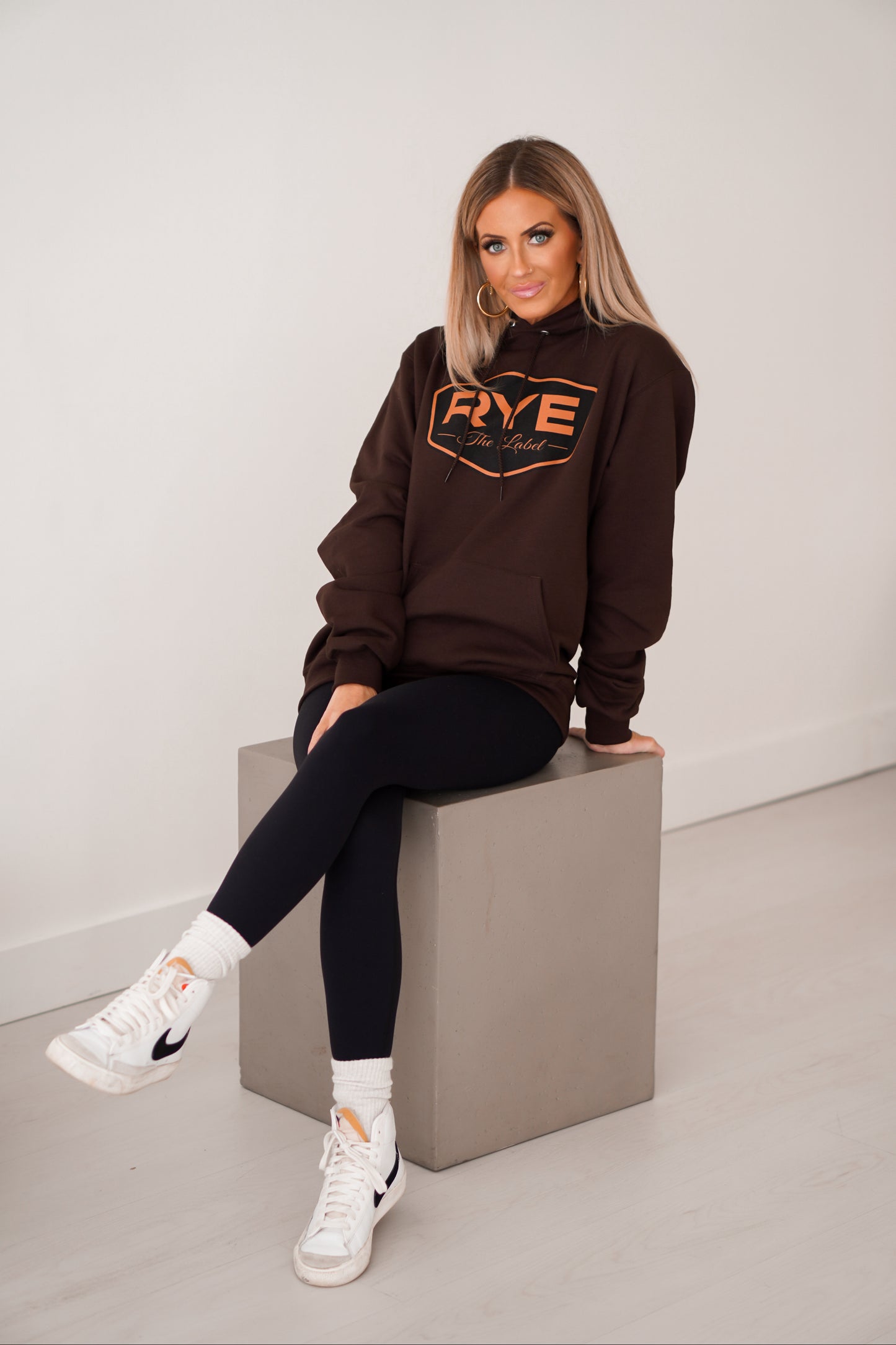 Girl sitting on gray box wearing brown RYE hoodie with legs crossed.