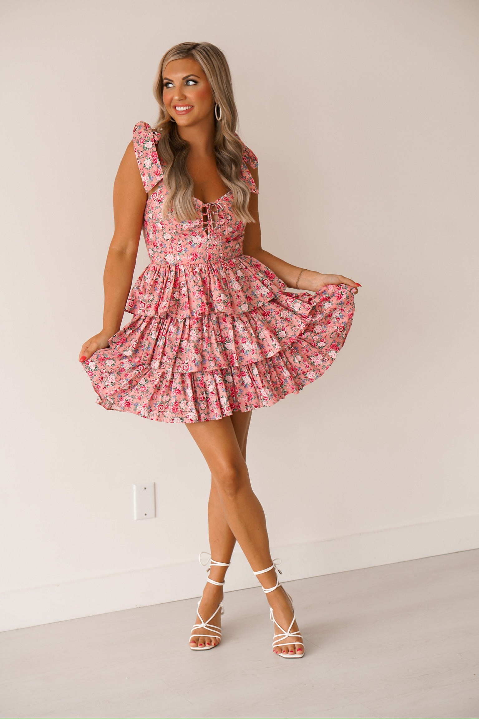 Blonde headed women standing against a white wall wearing a pink floral dress with white heels