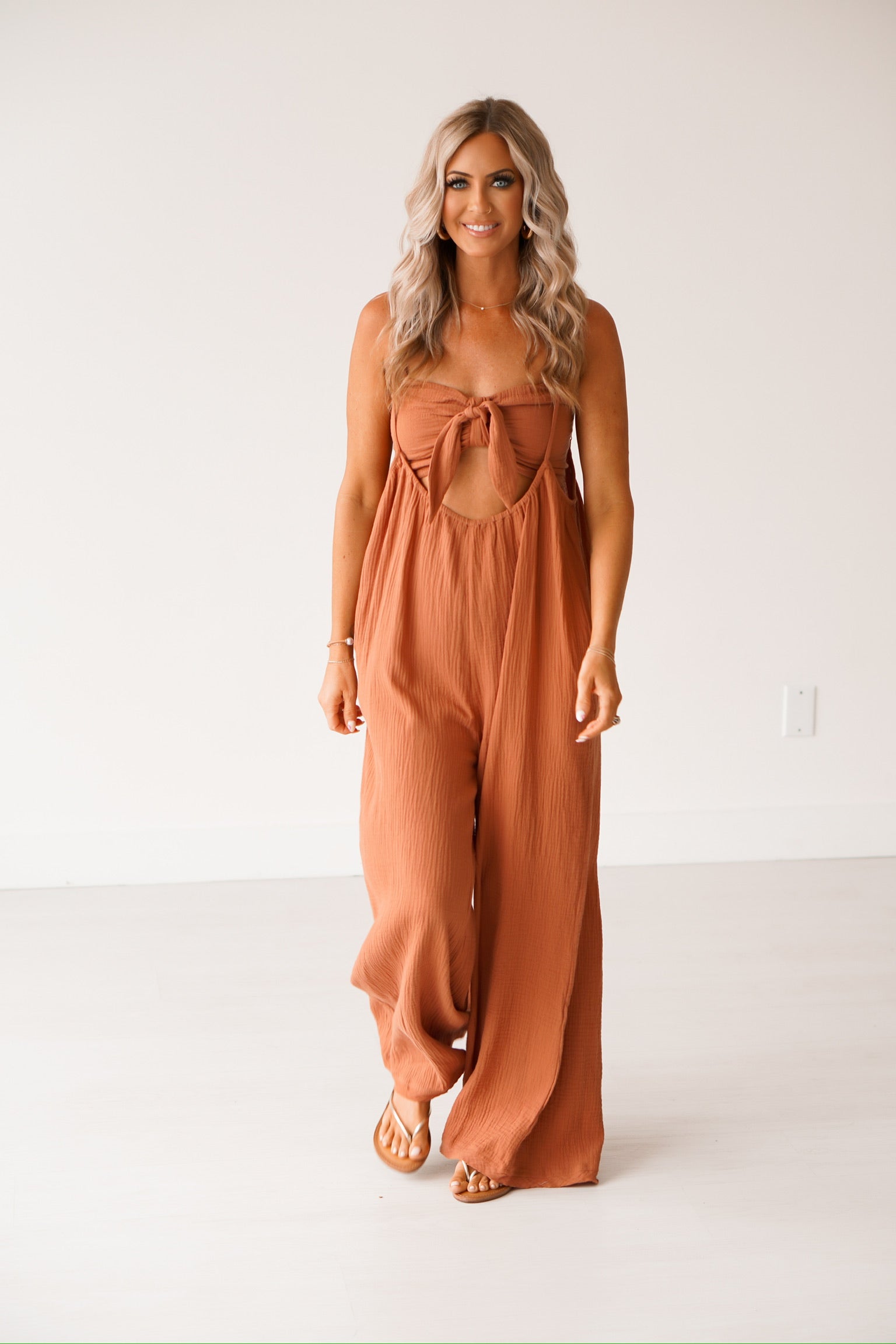 blonde headed lady wearing a brown jumpsuit against a white wall. It has a smocked top with adjustable straps and a bow on it