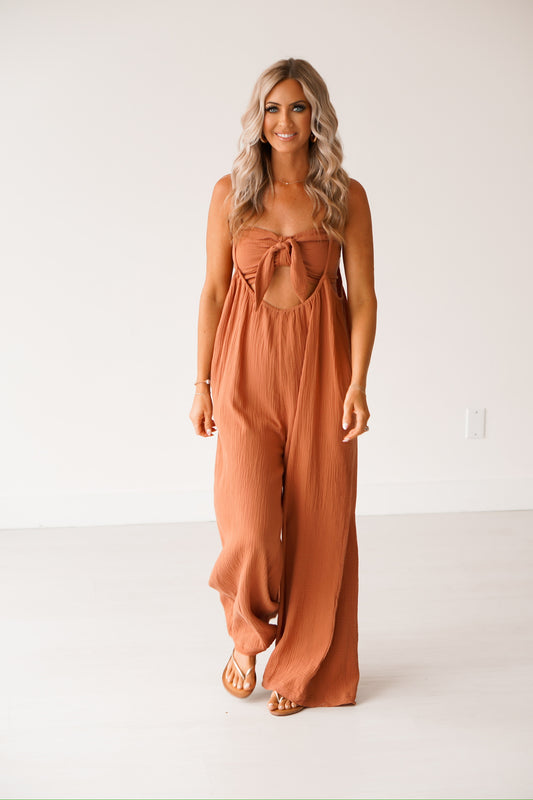 blonde headed lady wearing a brown jumpsuit against a white wall. It has a smocked top with adjustable straps and a bow on it