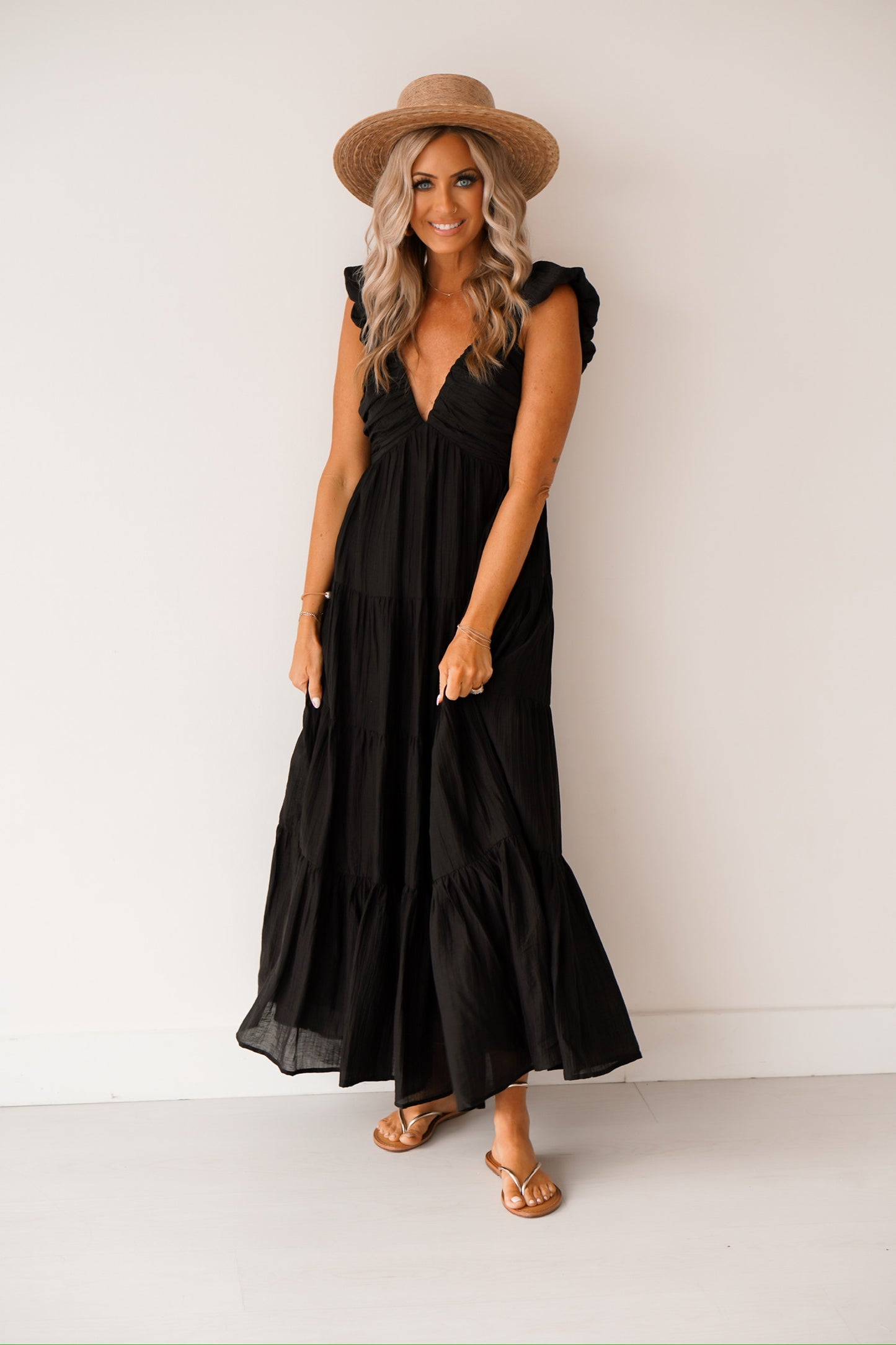 Blonde headed lady standing against a white wall wearing a black maxi dress with a hat 