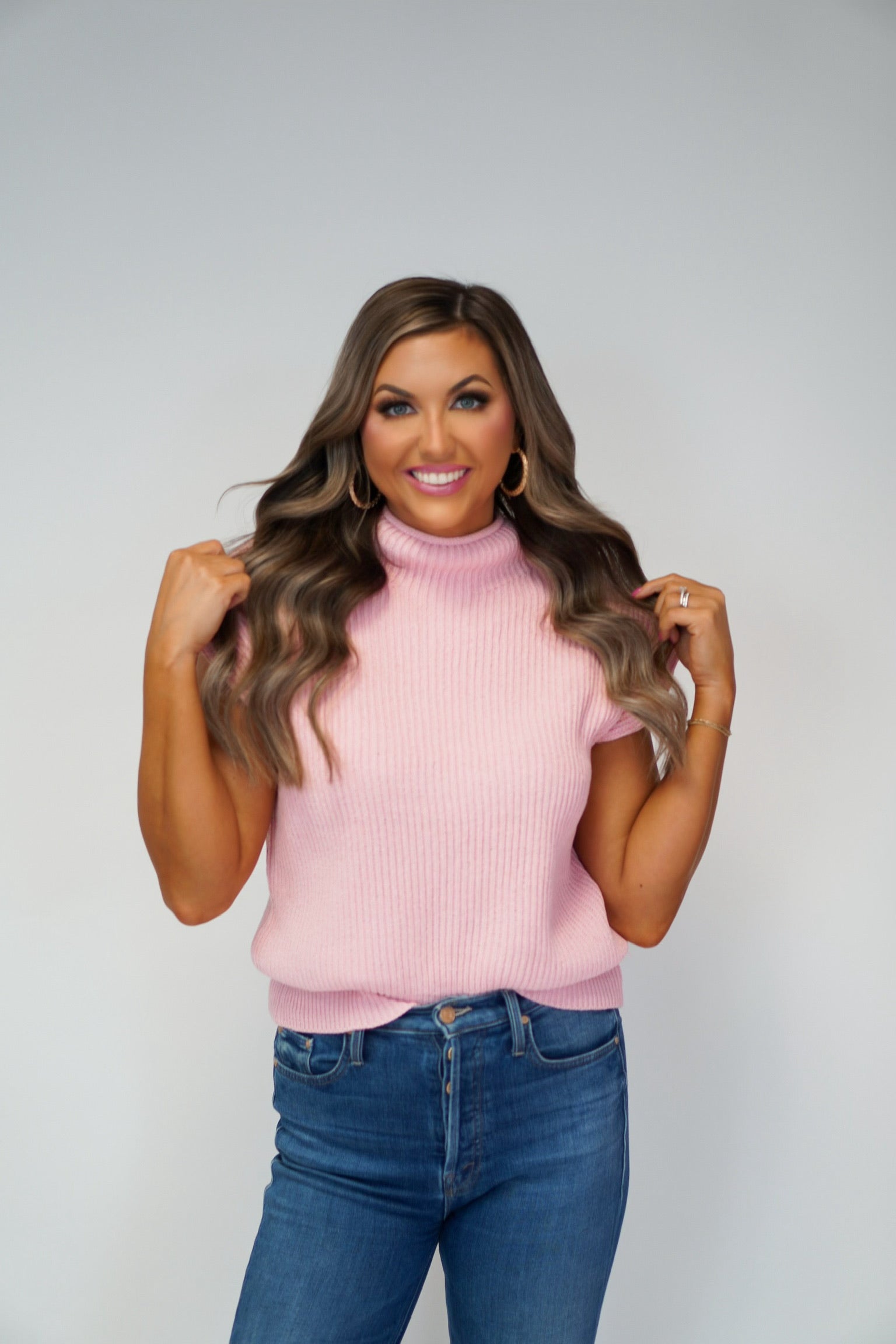 girl smiling playing with hair wearing a light pink mock neck sweater vest 