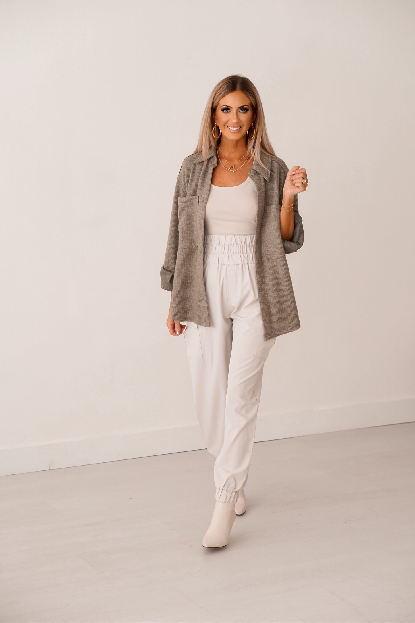 Blonde girl with cream tank, cream joggers with olive unbutton shacket. She is standing in front of white wall. 