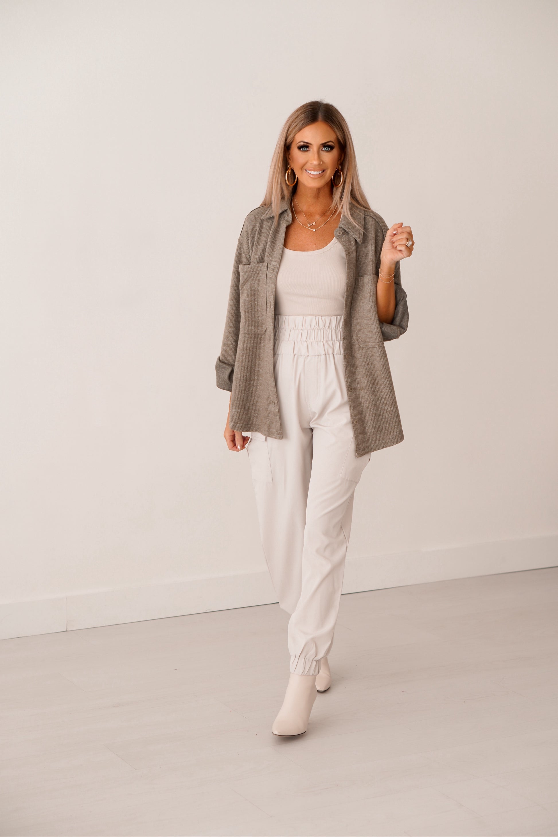Blonde girl with cream tank, cream joggers with olive unbutton shacket. She is standing in front of white wall. 