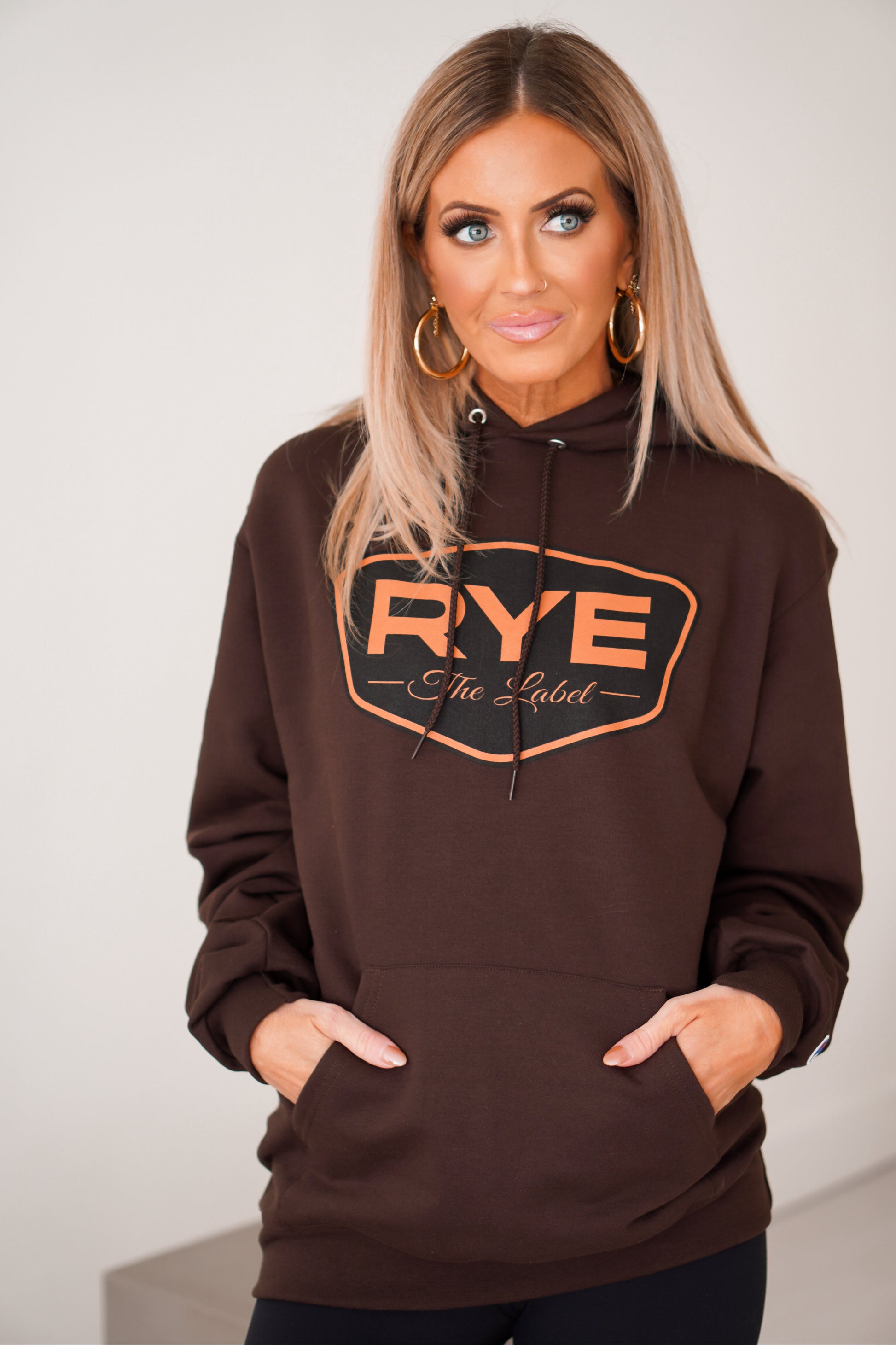Girl standing with brown RYE hoodie on with her hands in pockets.
