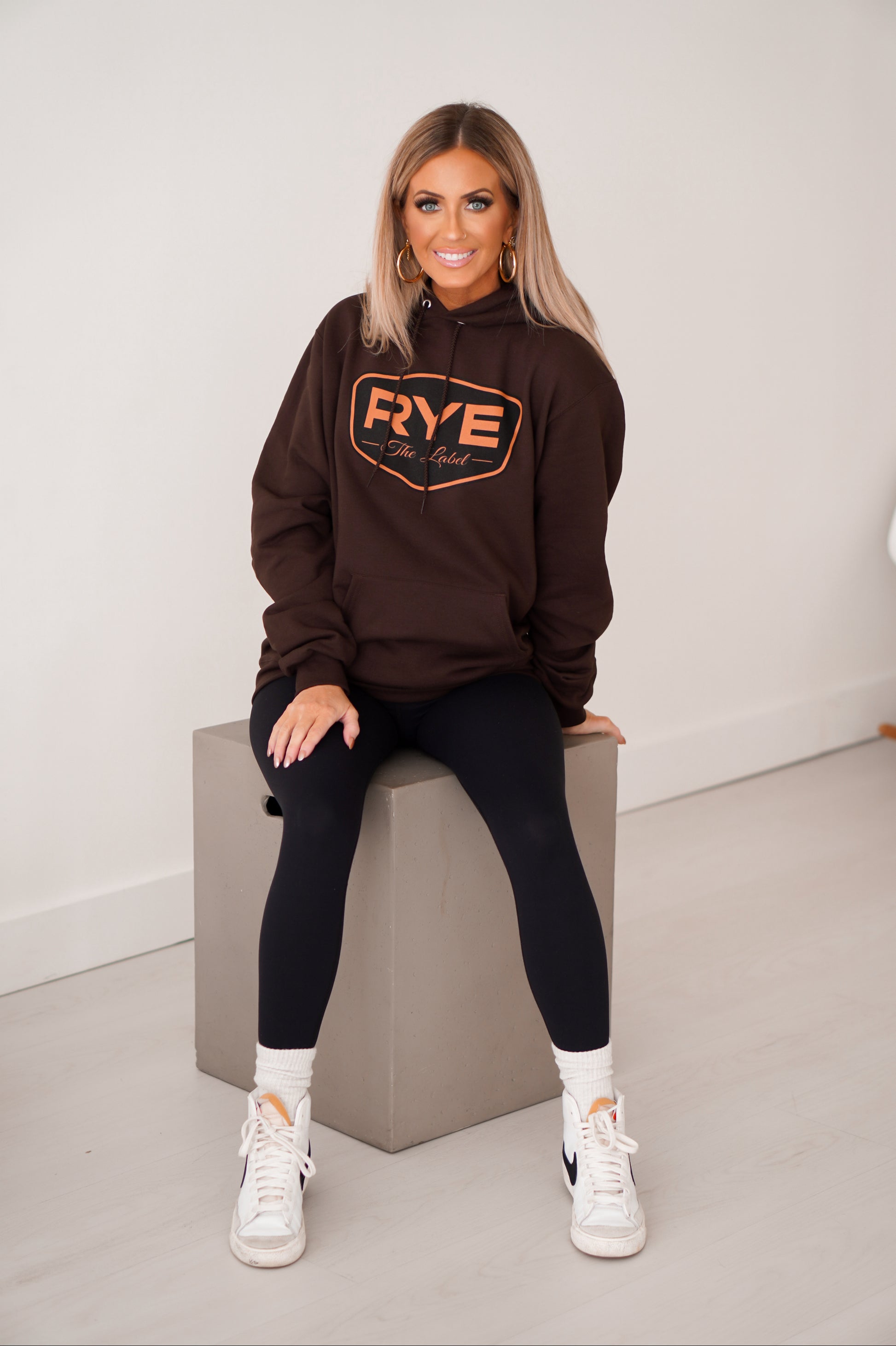 Girl sitting on gray box with a brown RYE hoodie on. 