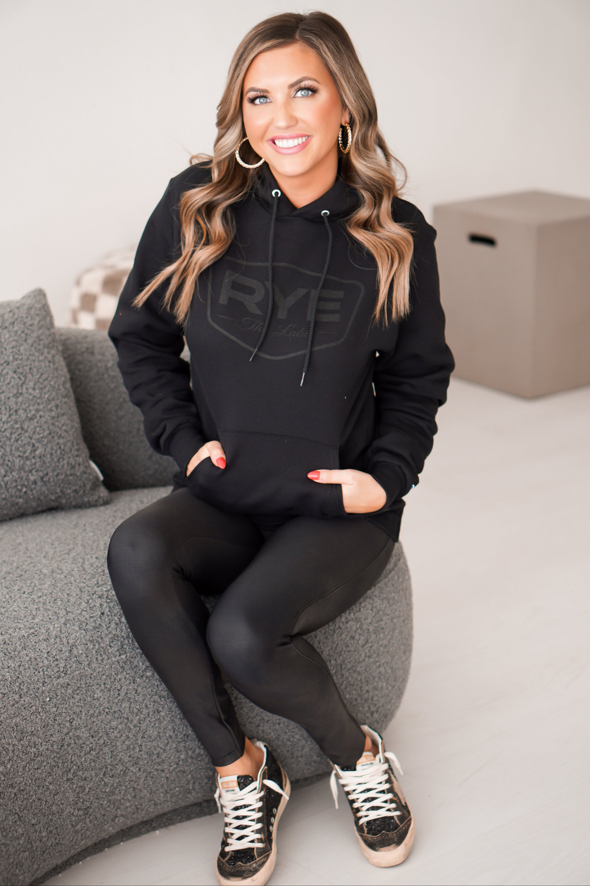 Girl sitting on gray couch with black RYE hoodie on with hands in pockets. 